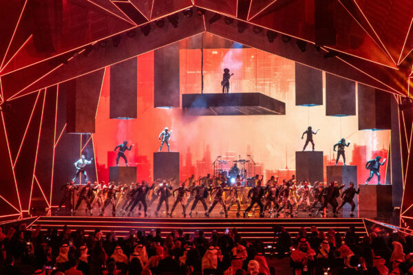 Image of stage performance with multiple dancers on stage, small number of dancers on podiums and guitarist on a raised platform