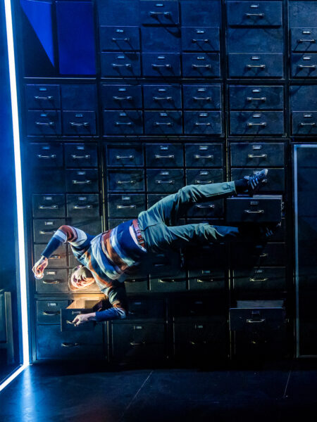 Photo of a man suspended on the face of a multi-drawer structure as part of a theatre performance